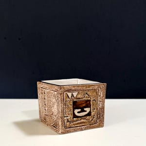 May include: A square, brown ceramic planter with a white interior. The planter features embossed geometric designs and a central square with a stylized letter. The planter is on a white surface with a dark blue background.