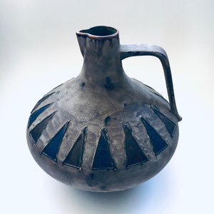 May include: A gray ceramic pitcher with a brown rim and a handle. The pitcher has a geometric design of black and blue triangles around the body.