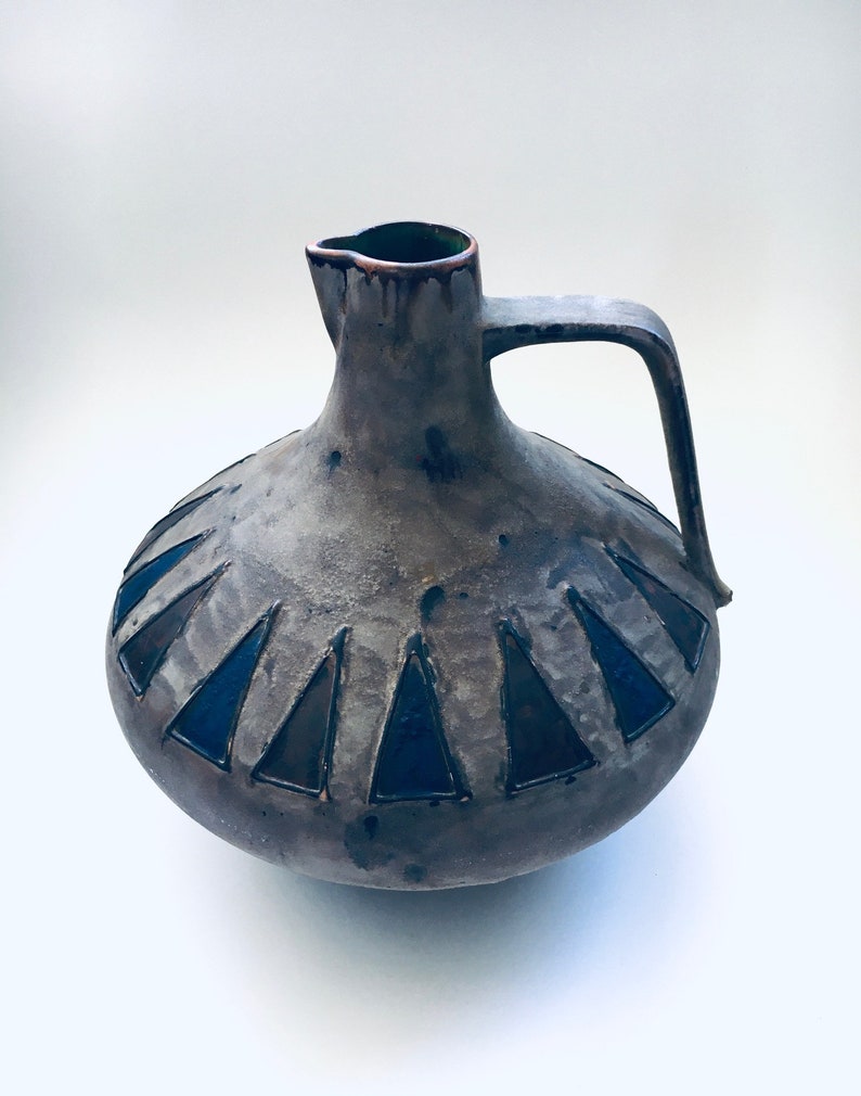 May include: A gray ceramic pitcher with a brown rim and a handle. The pitcher has a geometric design of black and blue triangles around the body.