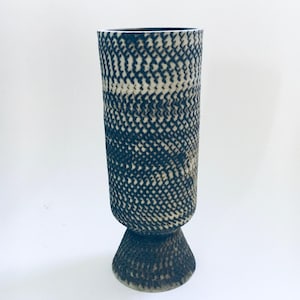 Könnte beinhalten: Eine hohe, zylindrische Vase mit einem strukturierten, kreuzschraffierten Muster in Dunkelblau und Creme. Die Vase steht auf einem ausgestellten, strukturierten Sockel. Das Keramikgefäß ist ein dekoratives Wohnaccessoire.