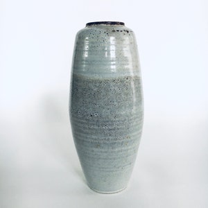 Puede incluir: Un jarrón alto de cerámica azul claro con un esmalte texturizado y moteado. El jarrón tiene una forma redondeada y cilíndrica y una abertura ligeramente acampanada.