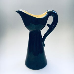 Puede incluir: Una jarra de cerámica negra con un interior de color crema y un asa curva. La jarra tiene una base alta y delgada y un pico ancho y redondeado.