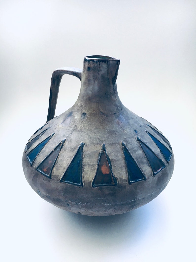 May include: A ceramic pitcher with a brown and gray glaze and a blue and black geometric design. The pitcher has a wide base and a narrow neck.