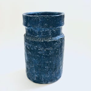May include: A cylindrical, dark blue ceramic vase with a textured surface. The vase has a slightly flared rim and a series of horizontal ridges around the middle. The overall aesthetic is rustic and handmade.