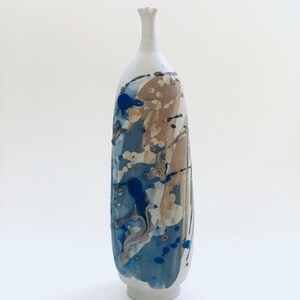 Può includere: Un vaso alto in ceramica bianca con un design astratto unico. Il vaso presenta schizzi di vernice blu, marrone e beige, creando un look dinamico e artistico.