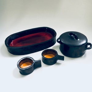 Könnte beinhalten: Ein Set aus vier schwarzen Keramik-Küchenartikeln. Das Set beinhaltet eine große ovale Servierplatte mit roter Glasur innen, eine kleine runde Schüssel mit gelber Glasur innen, eine etwas größere runde Schüssel mit gelber Glasur innen und einen kleinen schwarzen Topf mit Deckel.