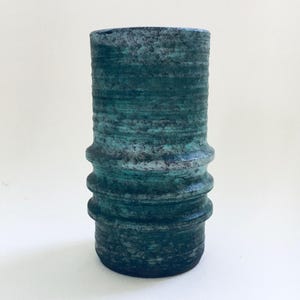 May include: A cylindrical, textured vase with a teal and blue-green color scheme. The vase has a series of horizontal ridges and a slightly flared base. The overall aesthetic is rustic and artistic.