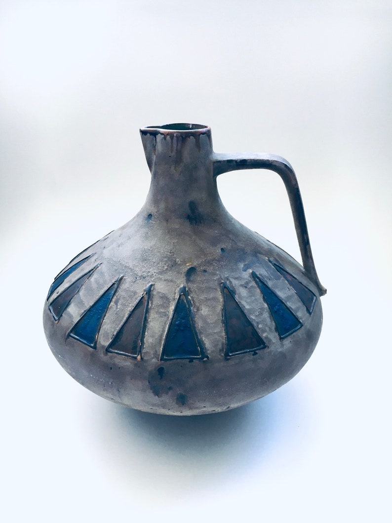 May include: A gray ceramic pitcher with a blue and brown geometric design. The pitcher has a wide base and a narrow neck with a handle.