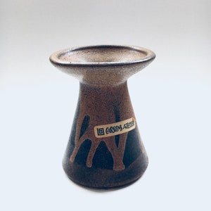 May include: A small, brown and black ceramic vase with a wide, flared top and a narrow base. The vase has a label that reads "Carstens Atelier".