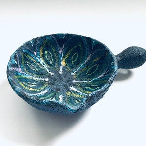 May include: A small, blue ceramic bowl with a textured surface and a handle. The bowl has a colorful, abstract design in shades of green, yellow, and white.