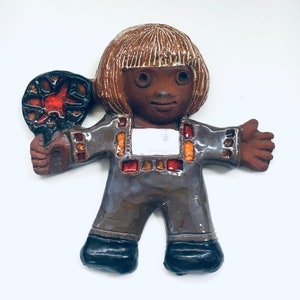 Puede incluir: Una figurita de cerámica de una persona con cabello castaño, piel morena y un atuendo colorido que sostiene una piruleta roja y negra. La figurita está de pie sobre un fondo blanco.