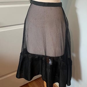 May include: Black mesh skirt with a ruffled hem, displayed on a wooden dress form. The skirt has a fitted waistband and a flared silhouette, showcasing a layered design. The sheer fabric creates a delicate, airy look.