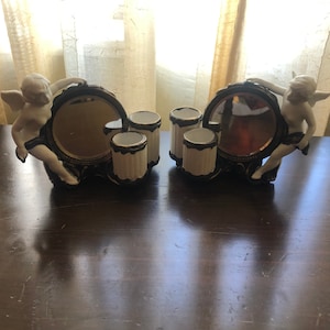 Puede incluir: Un conjunto de tocador vintage con dos figuras de querubines sosteniendo espejos redondos. Cada espejo tiene tres pequeños recipientes de cerámica blanca para guardar pinceles de maquillaje u otros artículos.