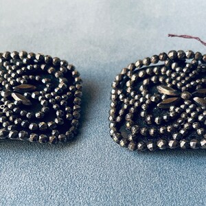 May include: A pair of antique silver-toned shoe buckles with a geometric design made of small, round beads. The buckles have a central design of two overlapping leaves.
