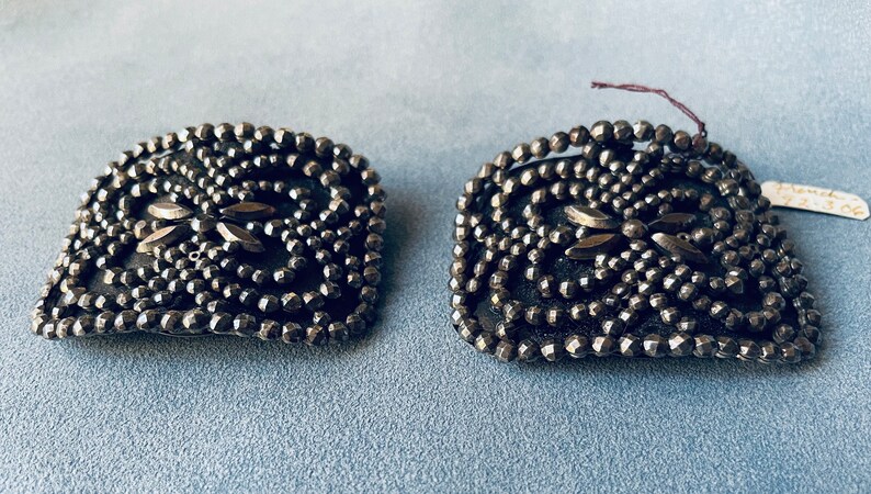 May include: A pair of antique silver-toned shoe buckles with a geometric design made of small, round beads. The buckles have a central design of two overlapping leaves.