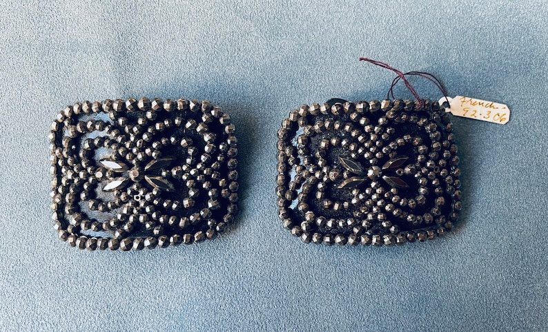 May include: A pair of silver-toned metal square-shaped brooches with a design of black beads and rhinestones. The brooches have a French tag attached to one of them that reads "French 92.306".