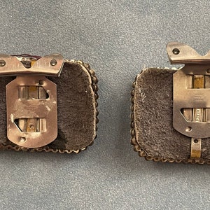 May include: Two silver metal clasps with a brown leather backing and a beaded border. The clasps have a metal hinge and a hook on the top.