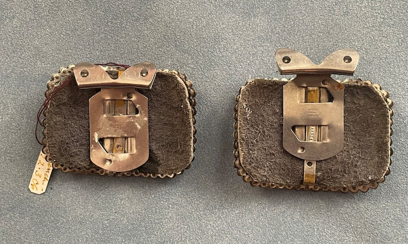 May include: Two silver metal clasps with a brown leather backing and a beaded border. The clasps have a metal hinge and a hook on the top.