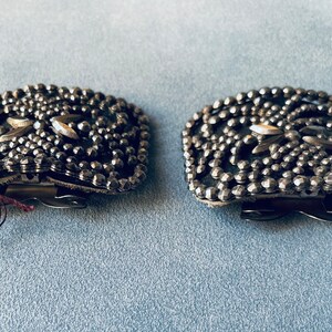 May include: A pair of silver-toned metal belt buckles with a decorative design of small, raised dots. The buckles have a square shape and a clasp closure.