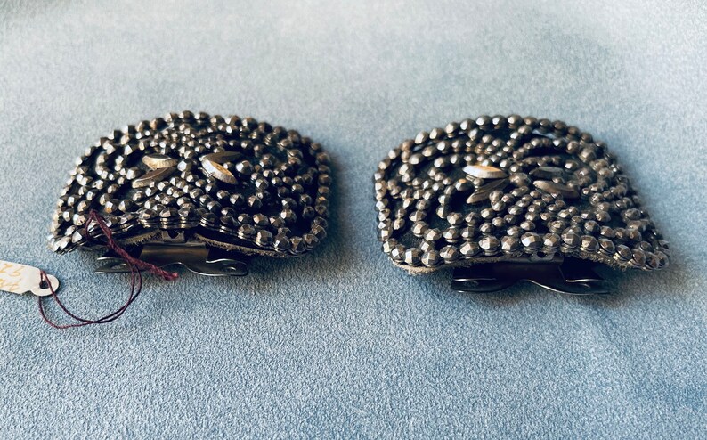 May include: A pair of silver-toned metal belt buckles with a decorative design of small, raised dots. The buckles have a square shape and a clasp closure.