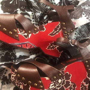 May include: A pair of brown leather wedge sandals with a red floral print and gold studs. The sandals have a strap across the toe and an ankle strap.