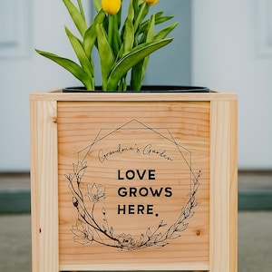 May include: Wooden planter box with yellow tulips. The box has a light wood finish and features the text "Grandma's Garden LOVE GROWS HERE" in a decorative design. The planter is a square shape with four legs.
