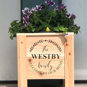 May include: A wooden planter box with the text "The Westby Family Welcome to Our Home" engraved on the front. The planter is decorated with a heart and a wreath of leaves. The planter is filled with purple and white flowers.