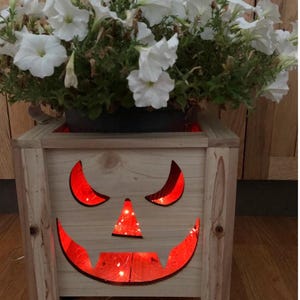 May include: A wooden planter box with a carved jack-o'-lantern face illuminated by red lights. The box holds a pot of white petunias. The planter is made of light-colored wood and has a rustic appearance, perfect for Halloween decor.