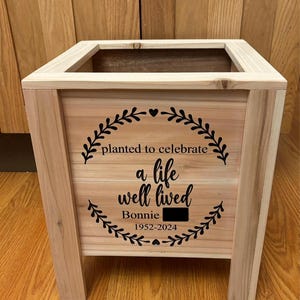 May include: A light-colored wooden planter box with a rectangular shape. The front of the planter features a black wreath design with the text "planted to celebrate a life well lived Bonnie 1952-2024". The planter has four legs and a rectangular opening at the top.