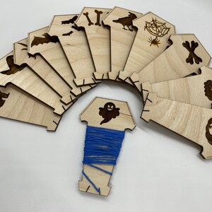 May include: A set of 12 wooden Halloween-themed embroidery hoops with a variety of spooky designs, including a cat, a ghost, a spider web, a skull, a pumpkin, and more. The hoops are arranged in a circle around a spool of blue thread.