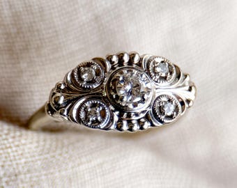 Pode incluir: Um anel de prata com um grande diamante redondo no centro e diamantes menores de cada lado. O anel tem um design detalhado com trabalhos de rolagem intrincados.