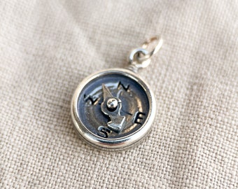 May include: A silver compass charm with a black face and a silver needle pointing north. The compass is on a light brown fabric background.