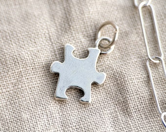 Puede incluir: Un colgante de pieza de rompecabezas de plata en una cadena de plata. El colgante es un diseño simple y clásico con un acabado liso.