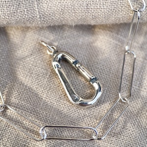 Peut inclure: Un collier en chaîne argent avec un pendentif en forme de mousqueton argenté. La chaîne est composée de maillons ovales.