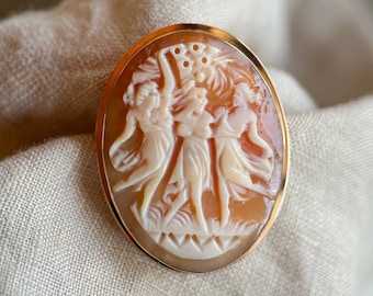 May include: A cameo brooch featuring a carved design of three women in a gold tone setting. The cameo is made of shell and has a light brown color.