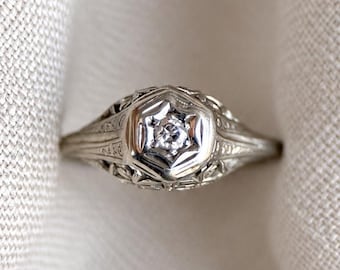 May include: A silver-toned metal ring featuring a central round clear gemstone set within a hexagonal frame. The band and setting display intricate filigree metalwork patterns, creating a detailed and textured appearance across the entire surface.