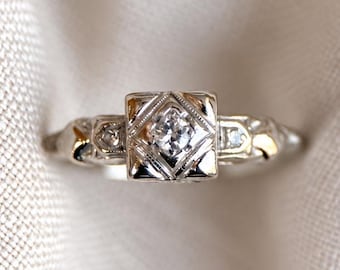 May include: A silver-toned ring featuring a central square setting with a single round clear stone. The band displays intricate metalwork with geometric patterns and smaller embedded stones on each side of the square centerpiece.