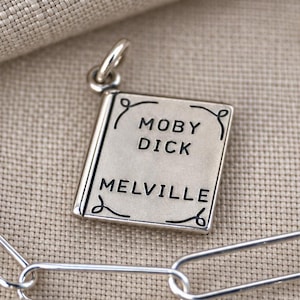 May include: A silver-toned metal charm shaped like a book, featuring the engraved text Moby Dick and Melville on the front cover, attached to a silver-toned paperclip chain. The charm includes decorative scrollwork lines at the corners.
