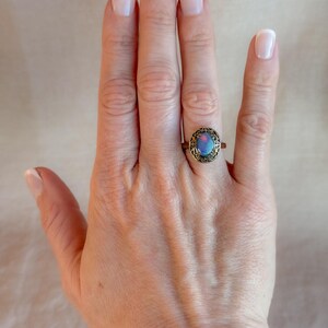 May include: A vintage-style ring with an oval, iridescent stone in shades of blue and purple. The ring has an ornate, bronze-colored setting. The ring is displayed on a hand with manicured nails.