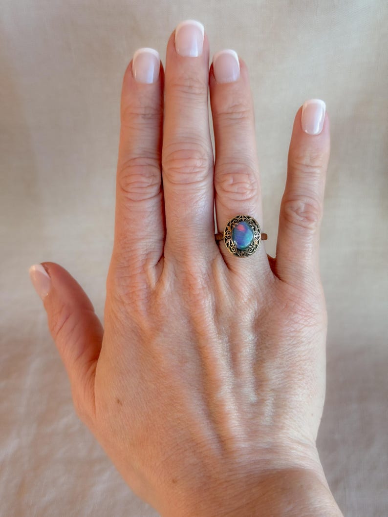 May include: A vintage-style ring with an oval, iridescent stone in shades of blue and purple. The ring has an ornate, bronze-colored setting. The ring is displayed on a hand with manicured nails.