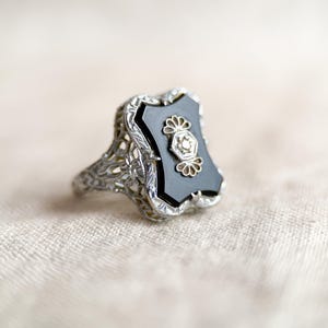 May include: A vintage silver ring with an onyx stone. The ring features an ornate filigree design on the band and around the black onyx stone. The onyx stone has a decorative floral design in the center.