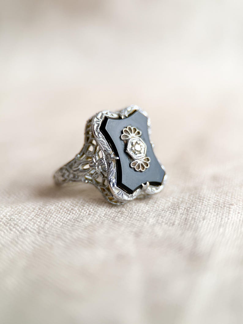 May include: A vintage silver ring with an onyx stone. The ring features an ornate filigree design on the band and around the black onyx stone. The onyx stone has a decorative floral design in the center.