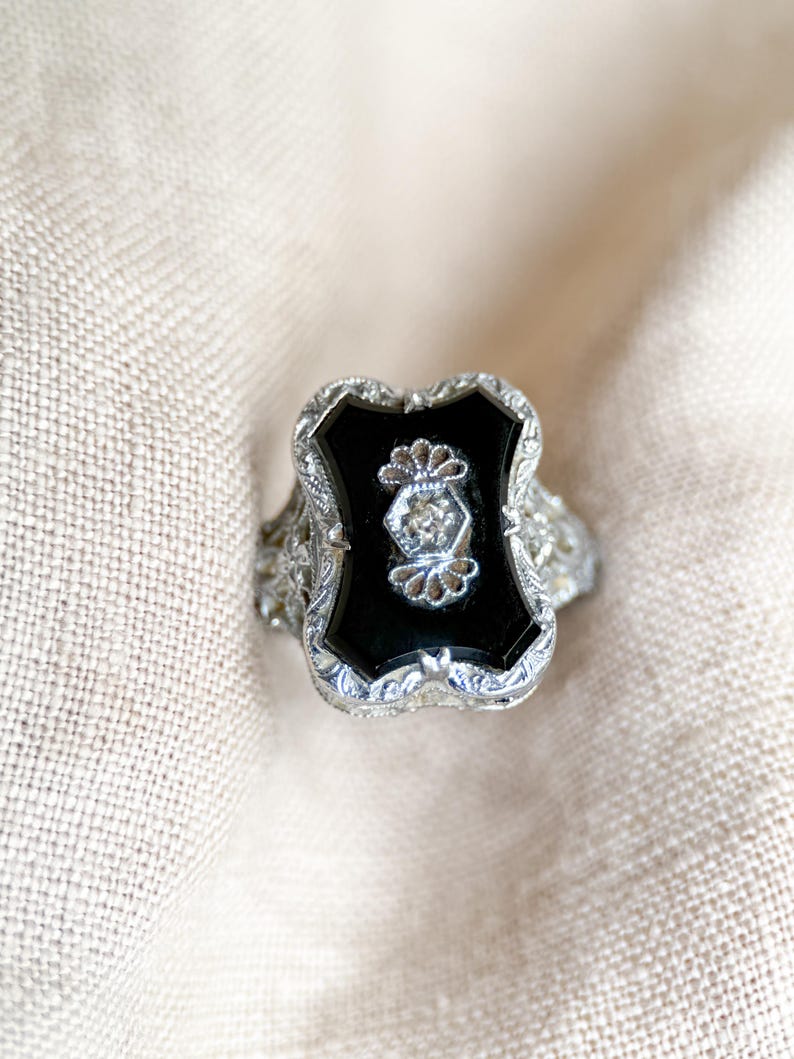 May include: An antique silver ring with a black onyx center stone. The ring features a shield-shaped onyx stone with a small diamond accent. The ring has intricate filigree detailing around the edges, creating a vintage aesthetic.