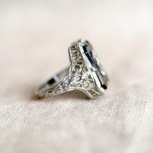 May include: A silver ring with an elongated, rectangular black gemstone. The ring features intricate filigree detailing around the gemstone and along the band. The ring is set against a soft, neutral background.
