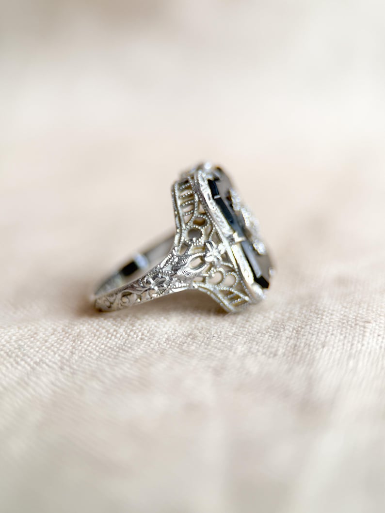 May include: A silver ring with an elongated, rectangular black gemstone. The ring features intricate filigree detailing around the gemstone and along the band. The ring is set against a soft, neutral background.