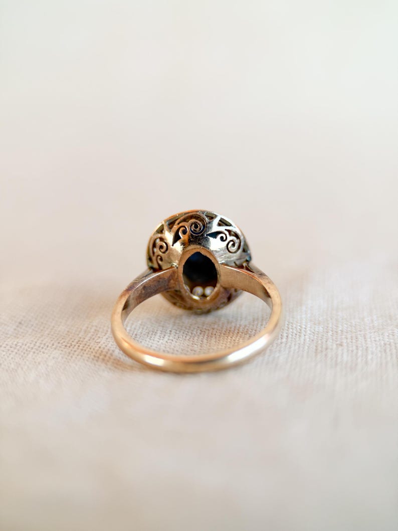 May include: A vintage gold ring with an oval, dark blue gemstone. The ring's setting features intricate filigree detailing around the gemstone. The band is a simple, polished gold. The ring is set against a neutral background.
