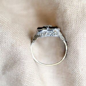 May include: A silver ring with an ornate filigree design. The ring features a rectangular black stone at the center, accented by a small, clear stone. The band is thin and delicate, set against a neutral fabric background.