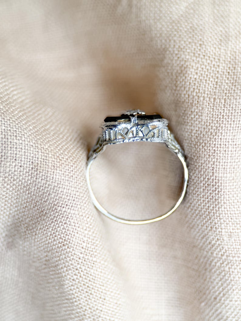 May include: A silver ring with an ornate filigree design. The ring features a rectangular black stone at the center, accented by a small, clear stone. The band is thin and delicate, set against a neutral fabric background.