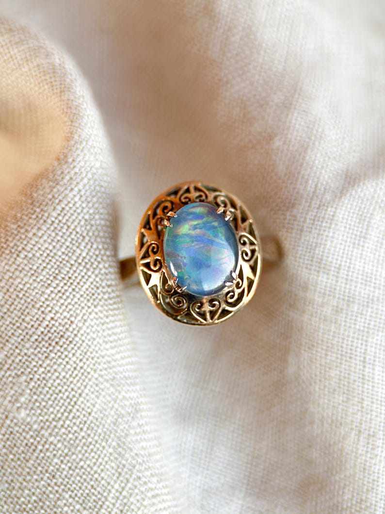 May include: A gold ring featuring an oval opal gemstone with iridescent blue and green hues. The opal is set within a filigree gold setting. The ring is displayed on a white fabric background.