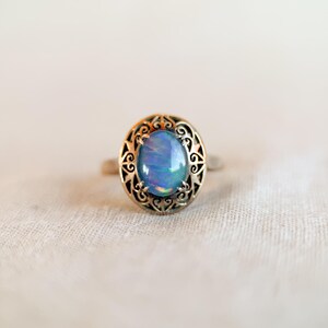 May include: A silver ring with an oval, iridescent blue gemstone. The gemstone is set in a decorative, filigree-style bezel. The ring is displayed on a white, textured surface, showcasing the jewelry's design and color.